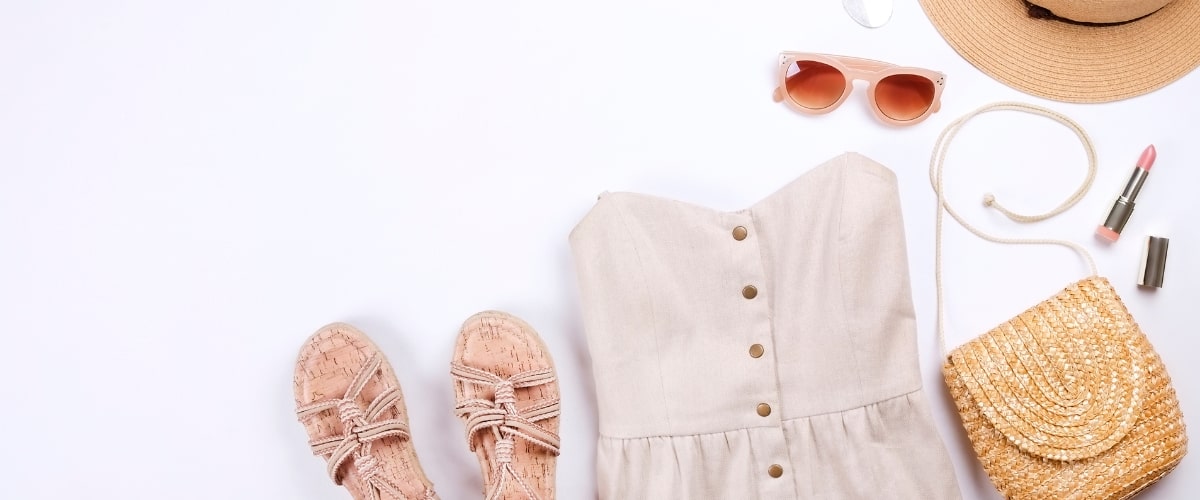 Flat lay of summer outfit for mature women featuring neutral button-down sundress, cork sandals, straw purse, sun hat, pink sunglasses, and lipstick on white background.