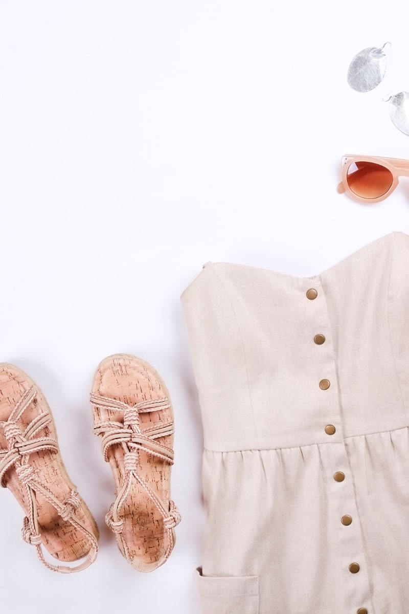 Flat lay of summer outfit for mature women featuring neutral button-down sundress, cork sandals, straw purse, sun hat, pink sunglasses, and lipstick on white background.