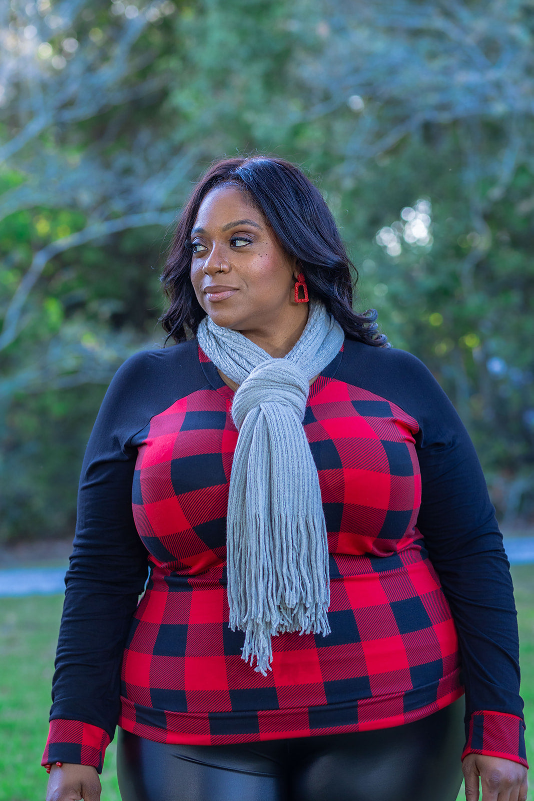 Raglan sleeve contrasting long sleeve top with red and black buffalo check print. This image features a gorgeous, plus size woman over 40 rocking this gingham print long sleeve crew neck top with a grey fringe scarf, red geometric earrings, and faux leather leggings from the fall collection at Jolie Vaughan Boutique.
