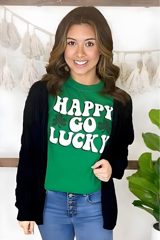 Smiling woman wearing a green St. Patrick's Day T-shirt with 'HAPPY GO LUCKY' text, styled with a black cardigan and blue jeans
